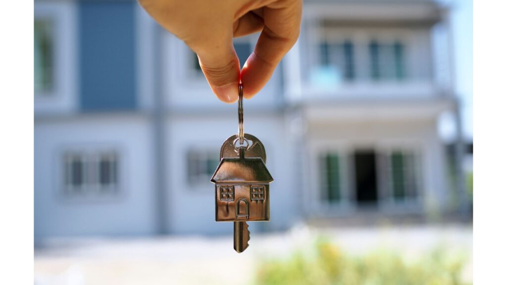 A hand holds a key with a keychain of a house. New home in the background